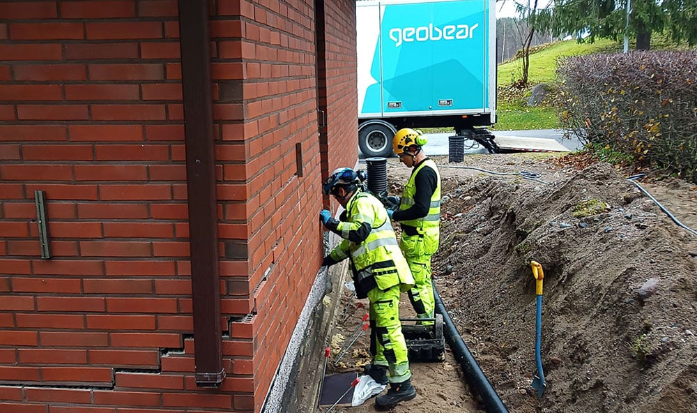 Två Geobear tekniker utför geopolymerinjektering på ett tegelhus. Halv Geobear lastbil syns i bakgrunden med loggan, markförstärkning villa.