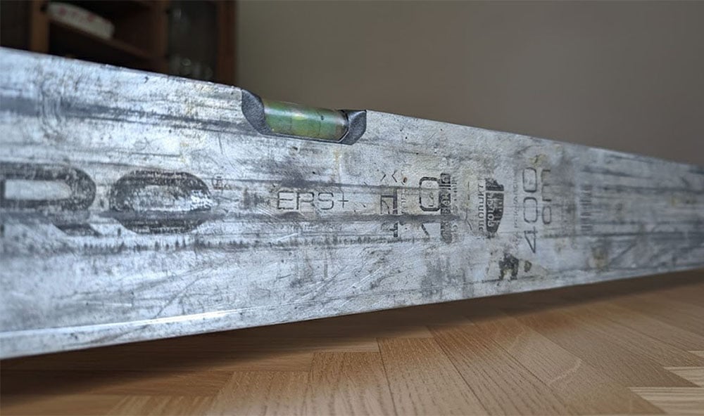 Ett silvrigt vattenpass ligger snett på fiskbensparkettgolv.